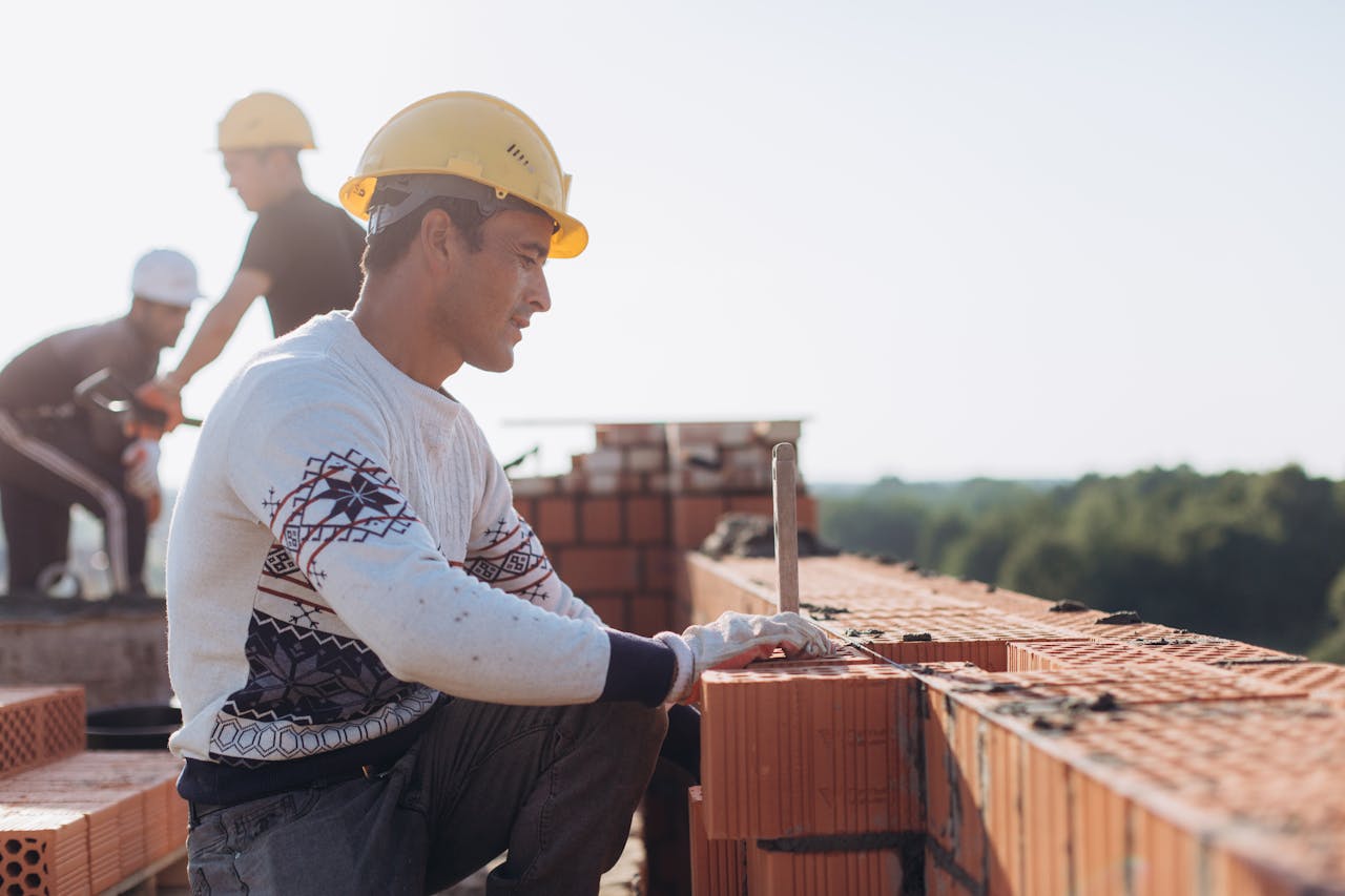Construction worker laying bricks outdoors in sunny weather with lush greenery in background.