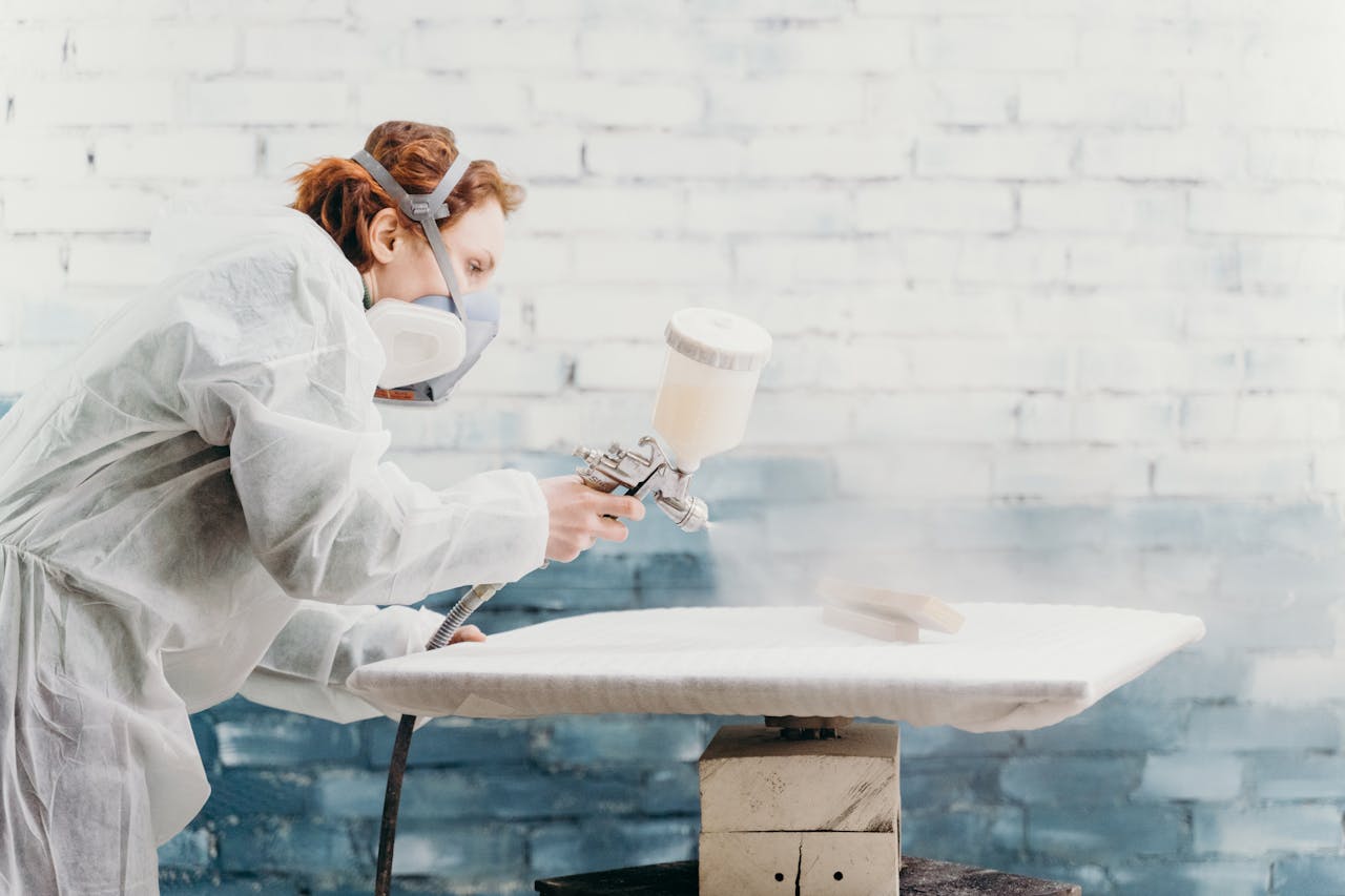 Redheaded woman wearing protective gear spray paints a furniture piece indoors.